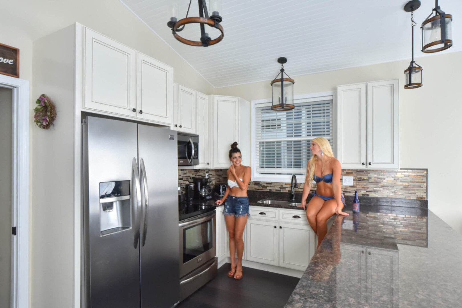 Two women in a swimsuit interacting in a white, fully equipped kitchen area with a dark brown marble counter top