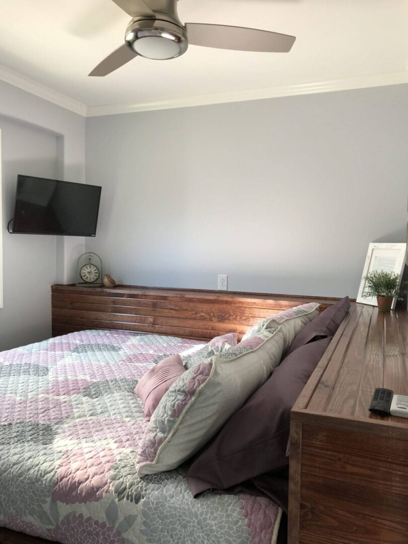 Light grey bedroom with a flat screen TV on the wall next to a dark brown full-length dresser bedframe of a bed