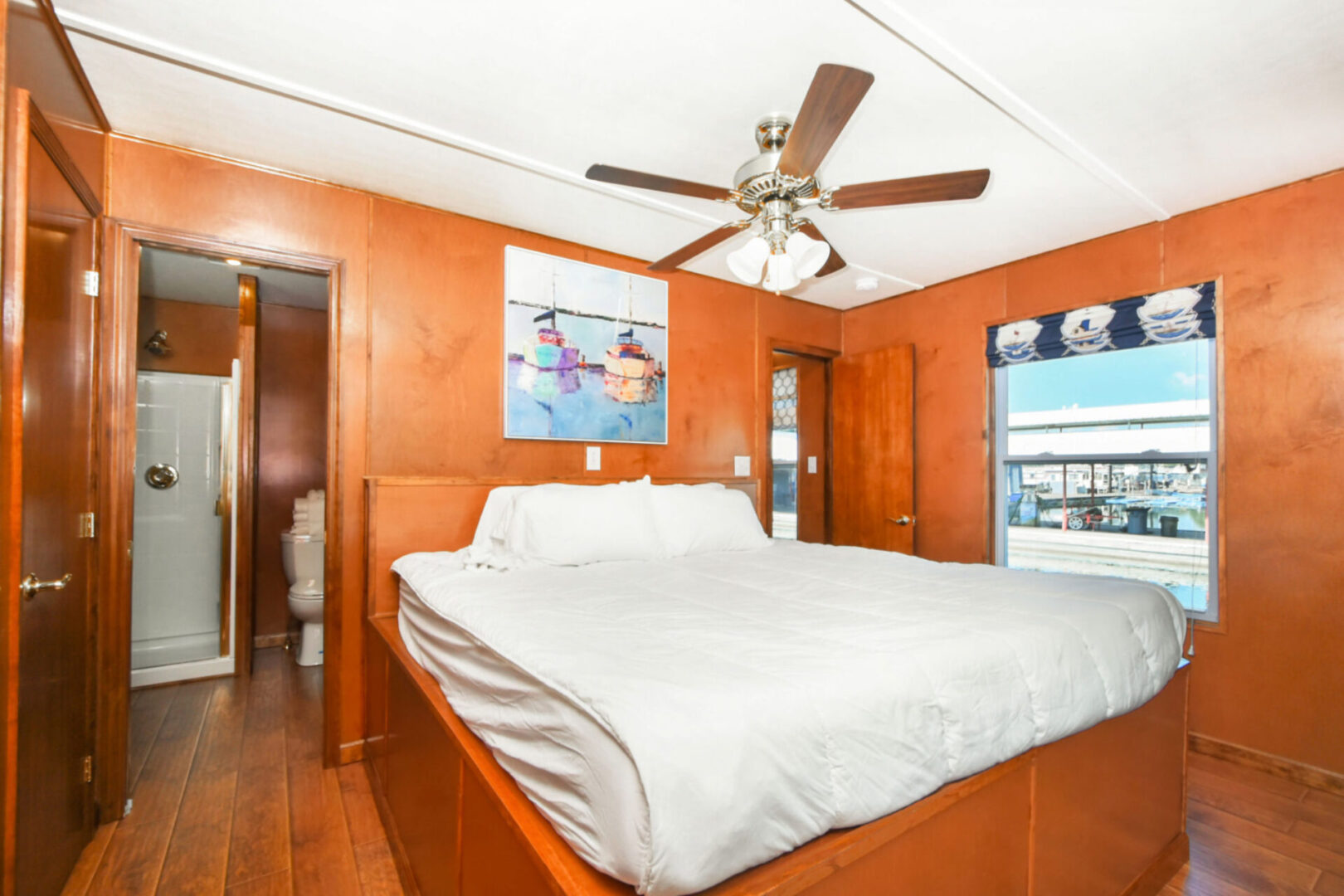 Boat artwork above the brown bedframe of a white bed inside a brown bedroom with a bathroom on to the left side